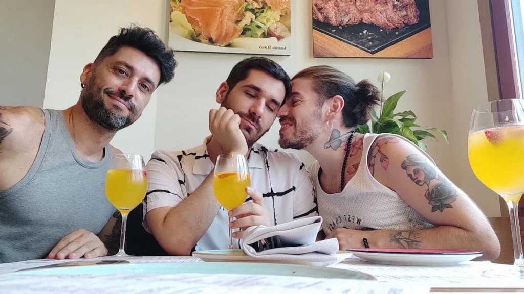 fotografia de una relación poliamorosa de tres hombres blancos con barba, sentados en un restaurante compartiendo unas mimosas naranjas.