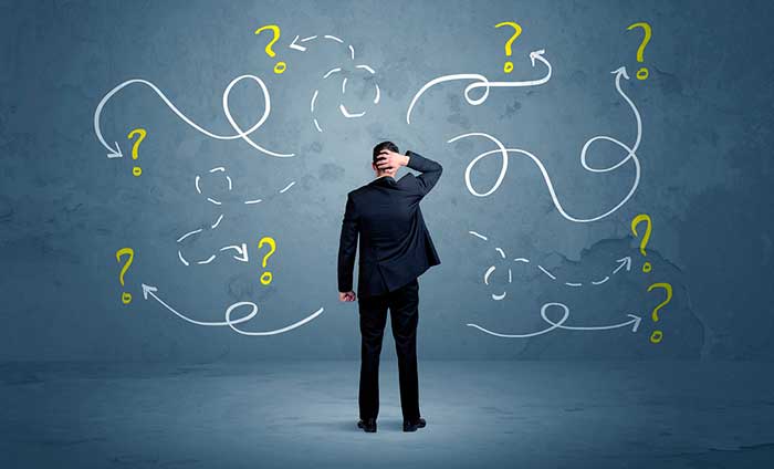 Hombre caucásico de espaldas con una mano en la cabeza y vestimenta de traje negro, preocupado por no saber cuál decisión tomar. Al fondo se aprecian signos de interrogación.